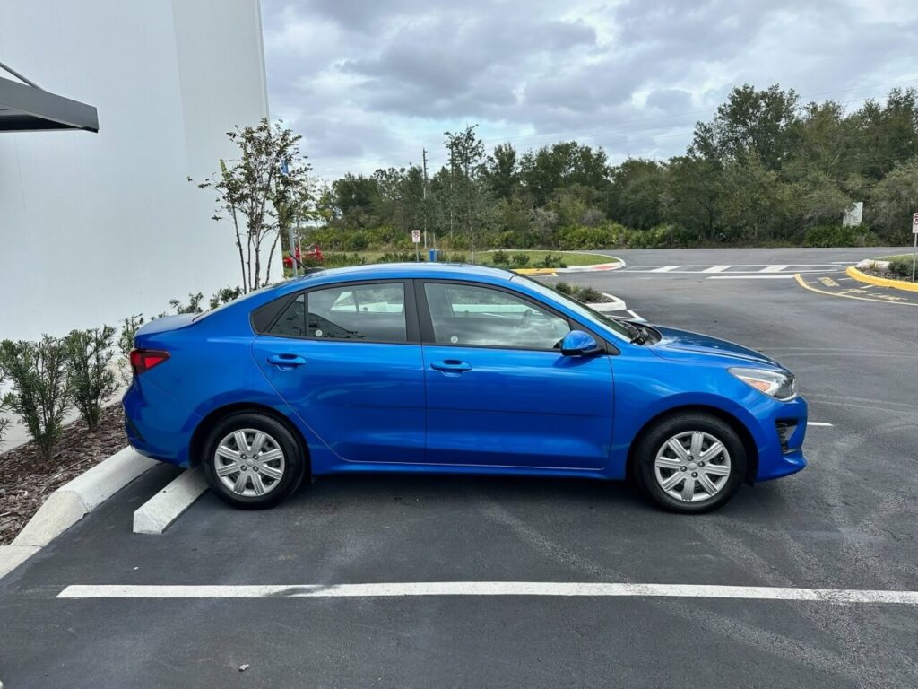 Sedan azul compacto da Move estacionado em vaga próxima a prédio branco, com fundo de árvores e céu nublado ao redor.